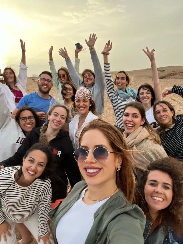 Un gruppo WeRoad sorride e alza le mani per un selfie in un ambiente sabbioso all'aperto al tramonto.