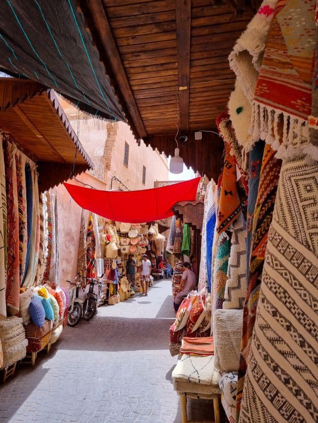 Una stretta via del mercato costeggiata da negozi che espongono tappeti colorati, stuoie e cesti sotto un tendalino di tessuto rosso.