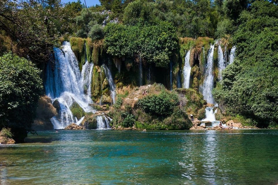 Ein mehrstufiger Wasserfall stürzt eine moosbewachsene Klippe hinab in einen türkisfarbenen Fluss, während sich Menschen auf den Felsen und im Wasser entspannen.