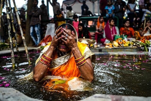 Una mujer con un sari amarillo y pulseras coloridas se cubre la cara mientras está sumergida en el agua durante una ceremonia religiosa.