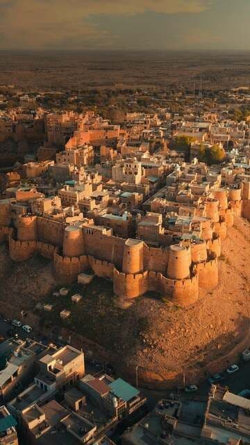 Una vista aérea de una gran ciudad fortificada de arenisca en una colina, con vistas a una ciudad moderna y un vasto paisaje desértico al atardecer.