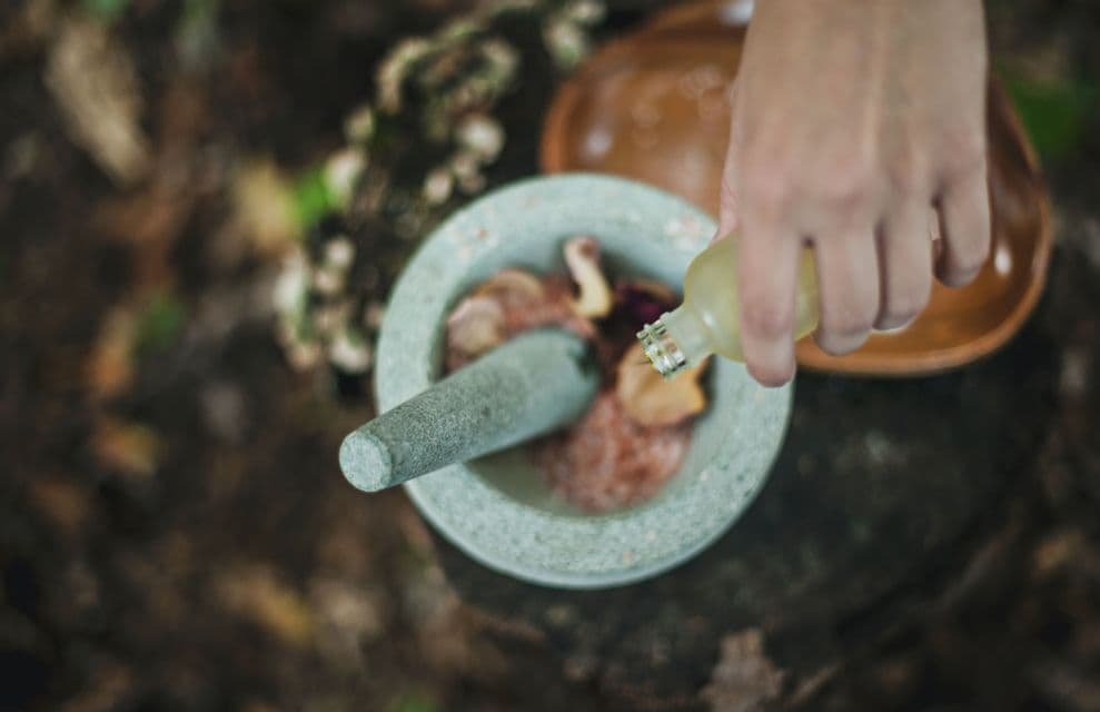 Vue de dessus d'une main versant du liquide d'une bouteille dans un mortier et un pilon en pierre avec des ingrédients naturels à l'intérieur.
