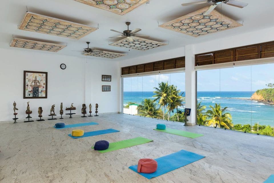 Un studio de yoga lumineux et vide avec des tapis et des coussins sur un sol en marbre, avec de grandes fenêtres donnant sur un océan tropical et des palmiers.