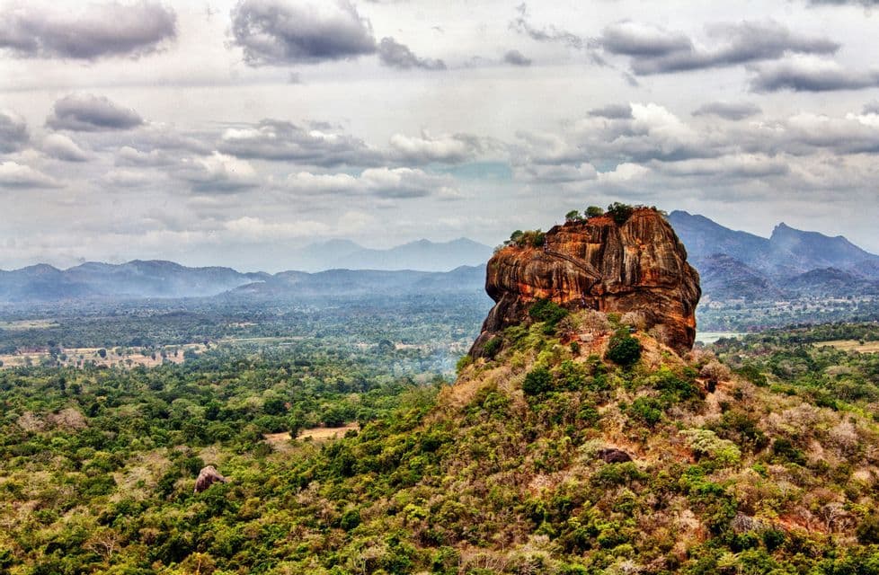 Une grande forteresse rocheuse brun-rougeâtre s'élève au-dessus d'un vaste paysage de forêt verte, avec des montagnes lointaines et brumeuses sous un ciel nuageux.