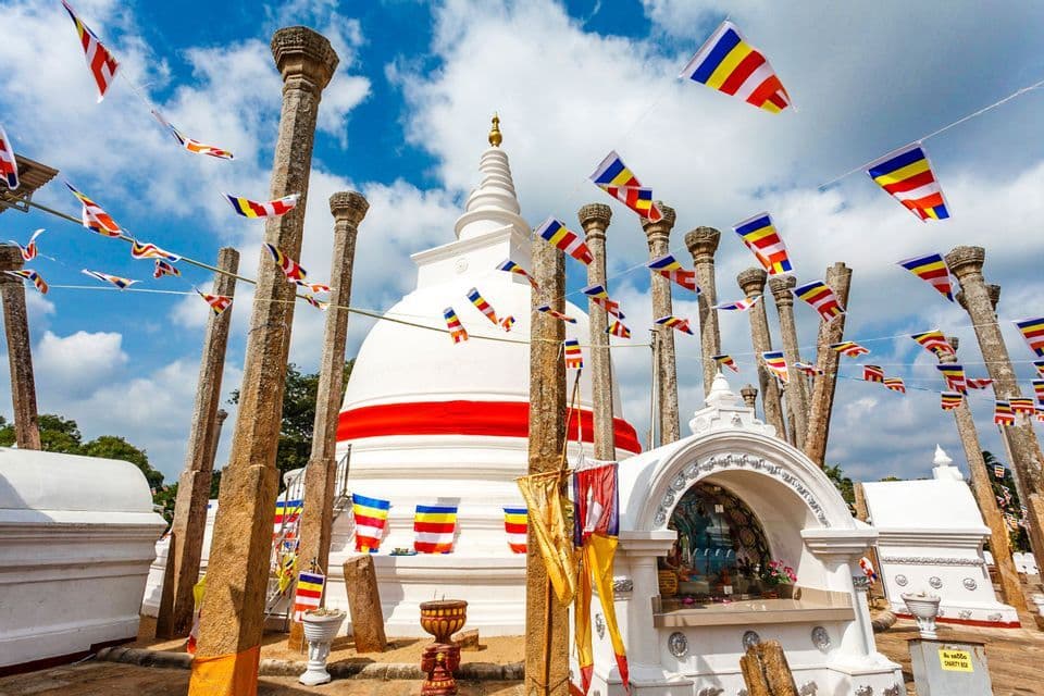 Un grand stupa blanc est entouré d'anciens piliers de pierre, avec des drapeaux bouddhistes colorés tendus au-dessus sous un ciel bleu nuageux.