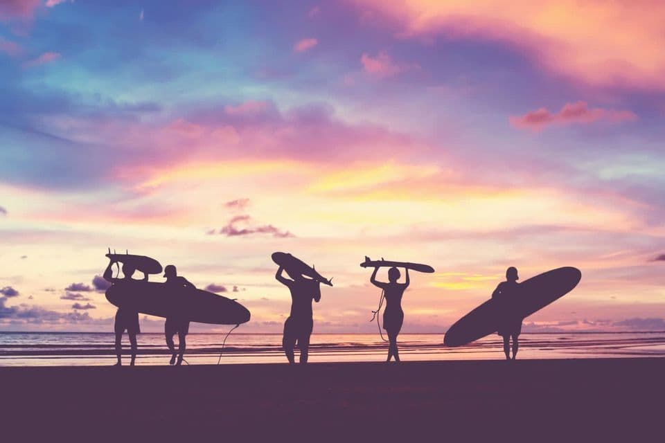 Silhouettes d'un groupe WeRoad avec planches de surf sur la plage au coucher du soleil.