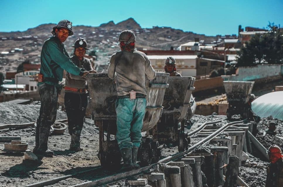 Un grupo de mineros con cascos y lámparas frontales trabaja con vagonetas en una vía férrea al aire libre.