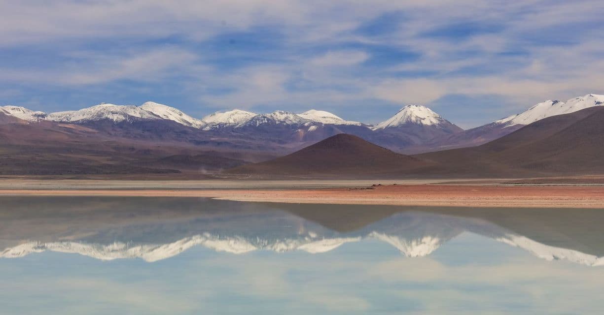 Una cadena de montañas cubiertas de nieve y colinas marrones se reflejan en las tranquilas aguas de un lago bajo un cielo azul con nubes ligeras.
