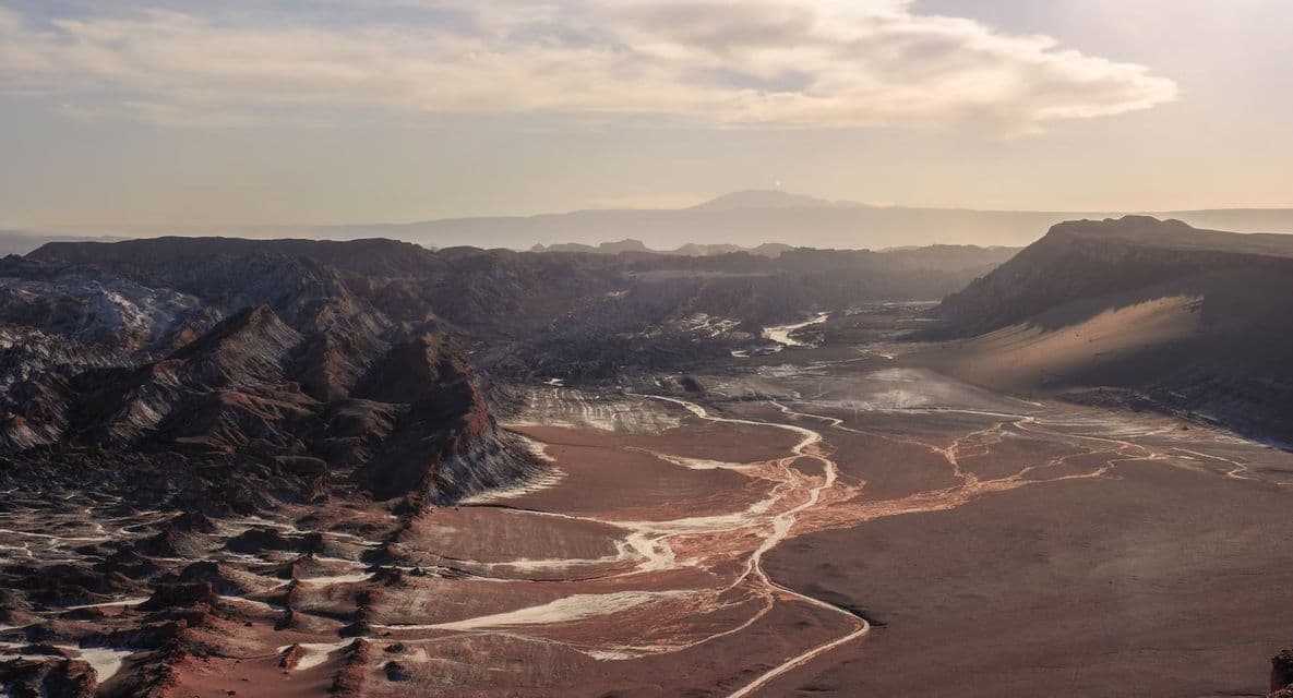 Un vasto valle desértico con oscuras montañas rocosas y sinuosos lechos de ríos secos de color claro bajo un cielo brumoso y luminoso.