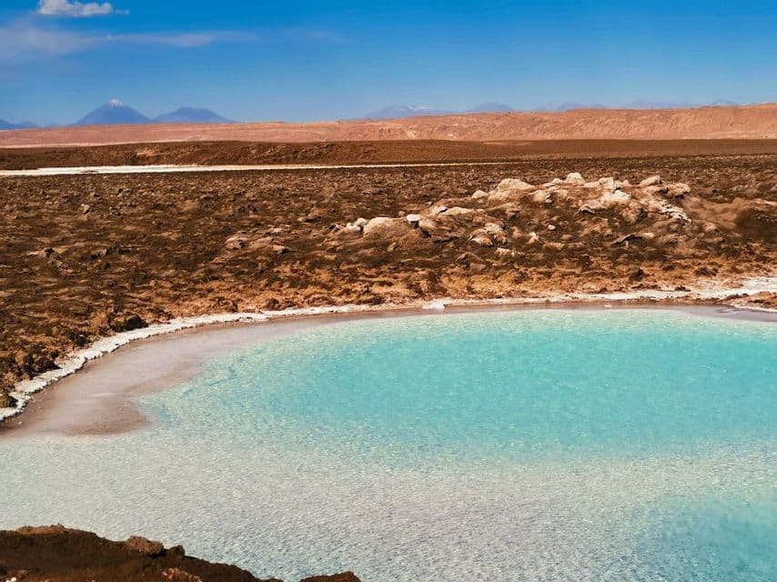 Una laguna turquesa con una orilla de sal blanca en un vasto desierto marrón, con montañas distantes visibles bajo un cielo azul.
