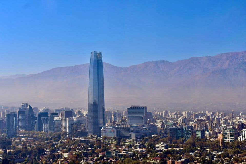 Un alto rascacielos de cristal domina un extenso horizonte urbano, con una cordillera montañosa difusa al fondo bajo un cielo despejado.