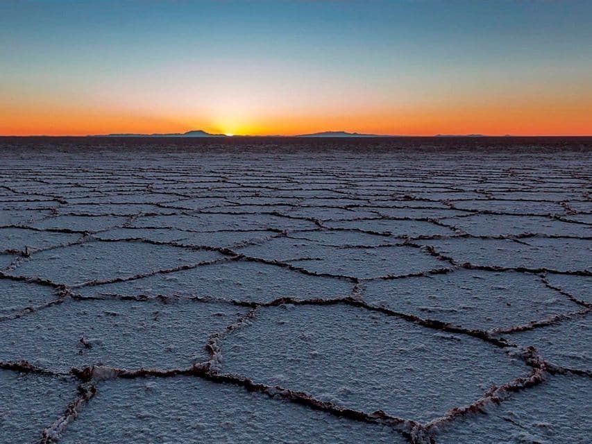 El sol se pone sobre un vasto salar agrietado, creando un resplandor naranja en el horizonte con montañas distantes.
