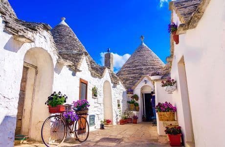 A bicycle with baskets of purple flowers is parked on a narrow street between whitewashed buildings with conical stone roofs under a blue sky.
