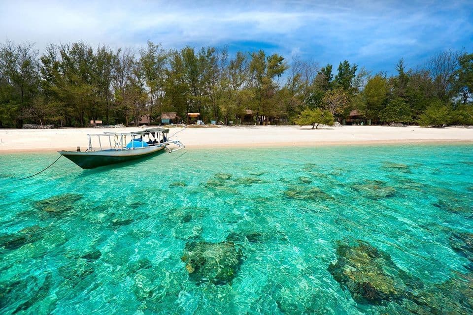 Un bateau flotte dans une eau turquoise peu profonde et transparente, révélant des rochers sur le fond marin, près d'une plage de sable bordée d'arbres.
