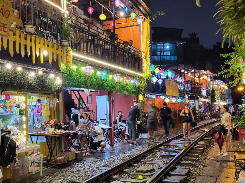 Un grupo de WeRoad camina por las vías del tren en una calle estrecha, bordeada de cafeterías y tiendas, e iluminada por coloridos farolillos por la noche.