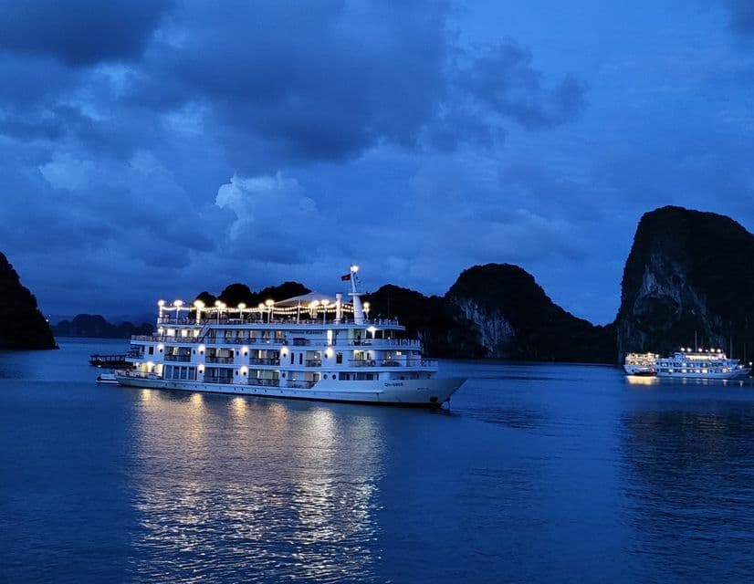 Un gran crucero con luces brillantes navega en aguas tranquilas al anochecer, con grandes formaciones rocosas al fondo.