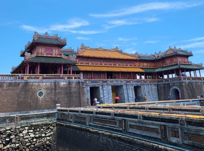 Un gran palacio asiático tradicional con tejados de tejas amarillas y verdes detrás de un puente de piedra y muros fortificados bajo un cielo azul.