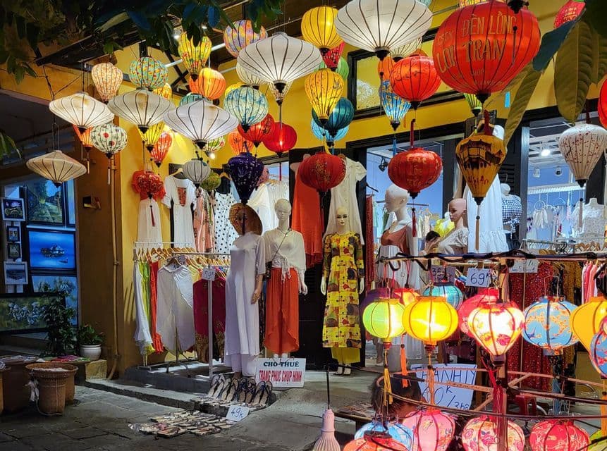 Farolillos de papel iluminados y coloridos cuelgan sobre la entrada de una tienda de ropa por la noche, con maniquíes vestidos con prendas tradicionales.