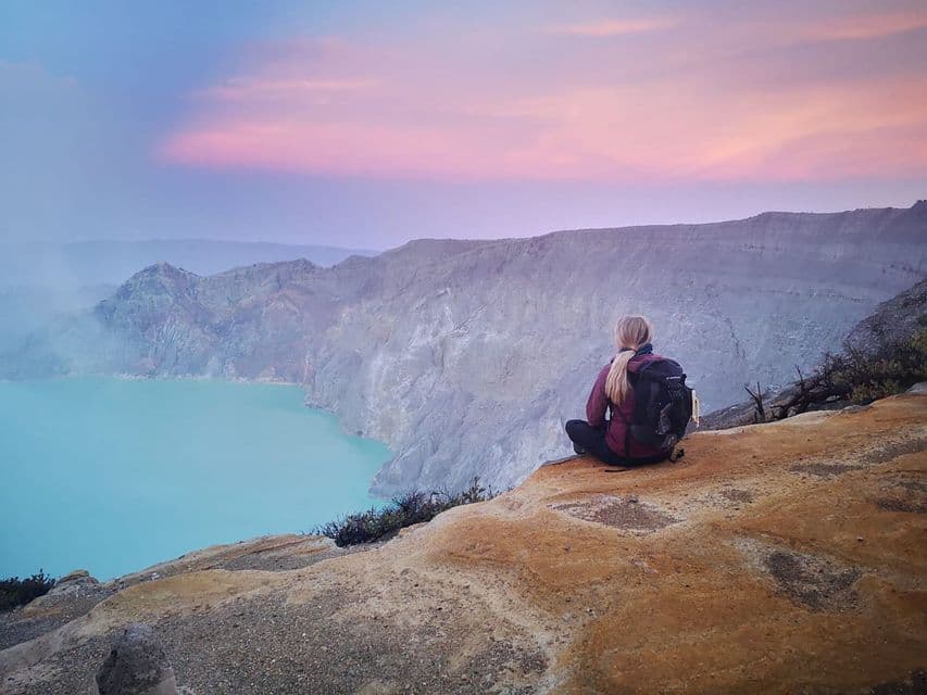 A person with a backpack sits on a rocky cliff edge, overlooking a turquoise crater lake under a pink and blue sky.