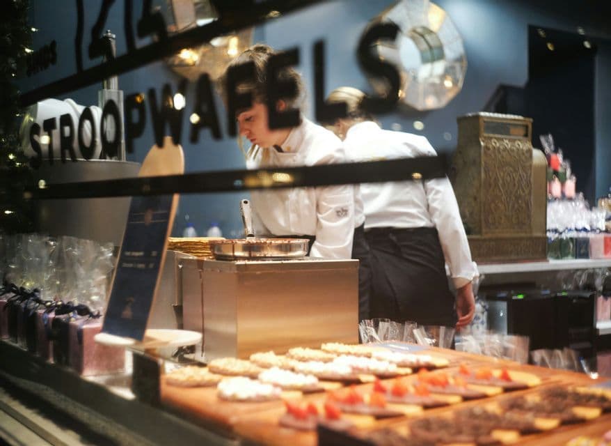 Un chef con uniforme blanco prepara stroopwafels frescos detrás del escaparate de una pastelería lleno de pasteles decorados.