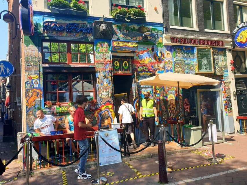 La fachada del coffeeshop 'The Bulldog', cubierta de murales coloridos, con gente sentada y de pie afuera en un día soleado.