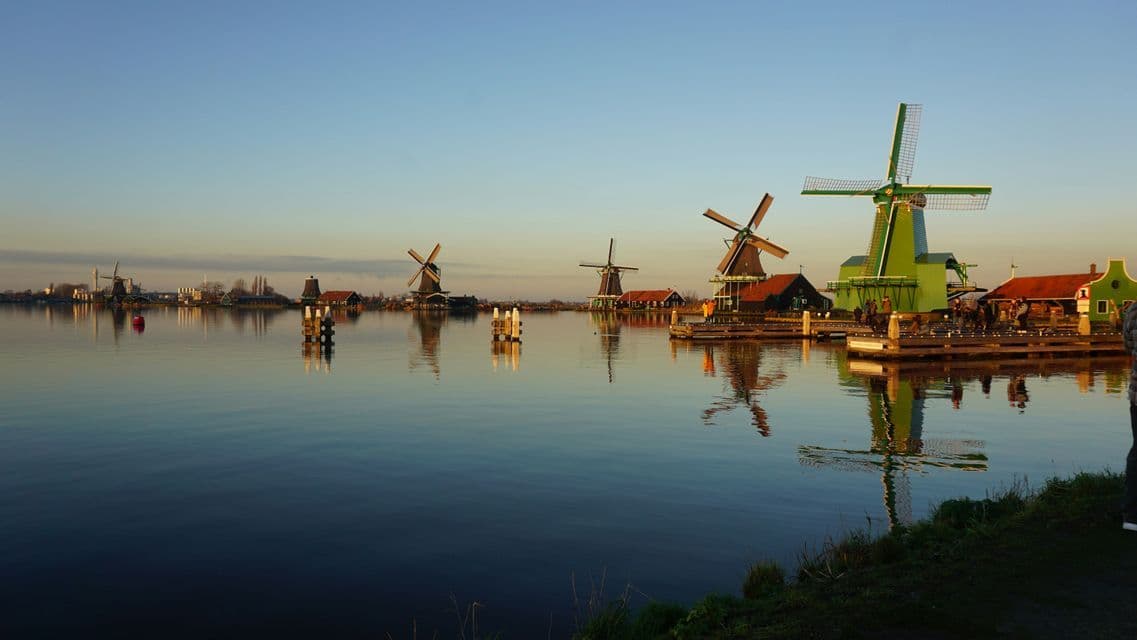 Un grupo de WeRoad en un muelle observa molinos de viento tradicionales reflejados en un río tranquilo al atardecer.