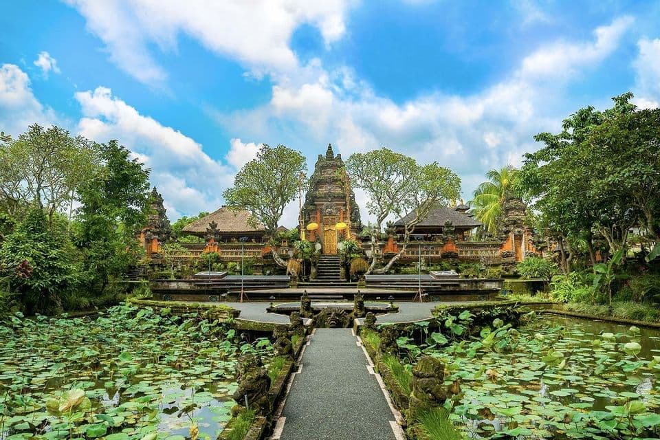 Ein steinerner Pfad führt über einen mit Seerosen bedeckten Teich zu einem verzierten Tempel, umgeben von Bäumen unter einem blau-weißen Himmel.