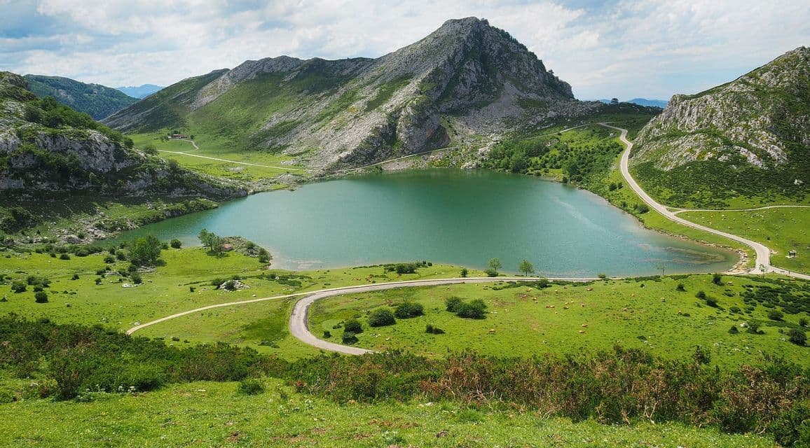 Un camino sinuoso bordea un lago turquesa en un valle rodeado de colinas verdes y una montaña rocosa bajo un cielo parcialmente nublado.