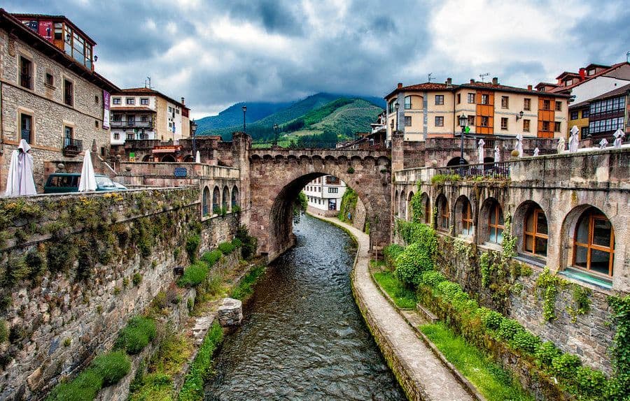Un puente de arco de piedra cruza un río que fluye a través de un pueblo histórico, con montañas verdes al fondo bajo un cielo nublado.