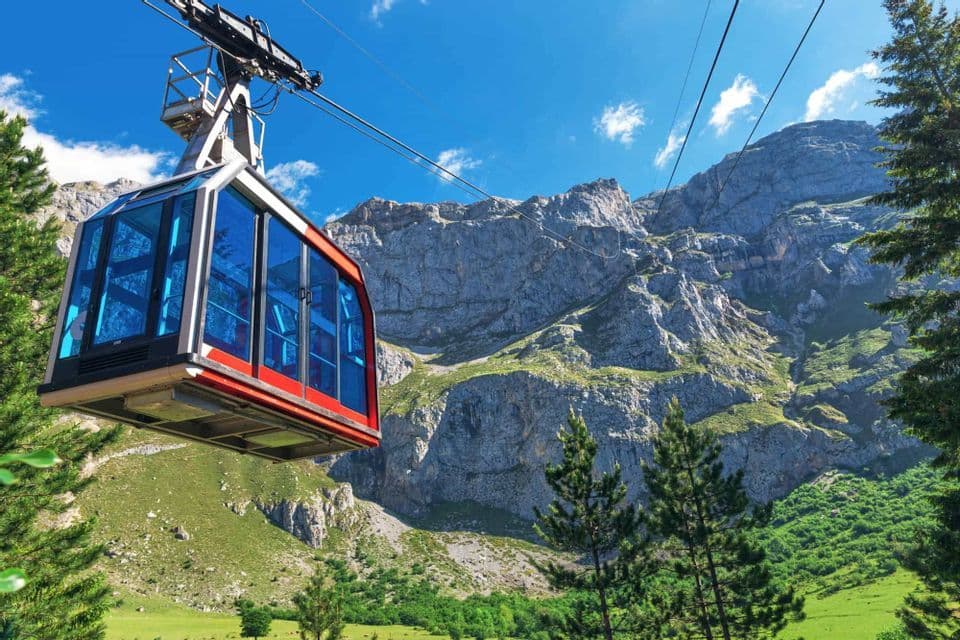 Un teleférico rojo con ventanas azules asciende sobre un valle de montaña verde bajo un cielo azul claro.