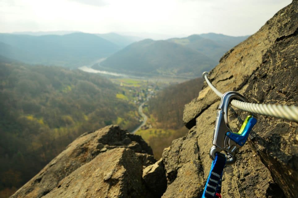 Un mosquetón sujeto a un cable de seguridad de acero en una vía ferrata, en lo alto de un acantilado con vistas a un valle de montaña.