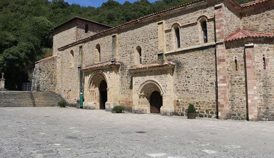 Un histórico monasterio de piedra con arcos se alza en un gran patio empedrado al pie de una colina verde.