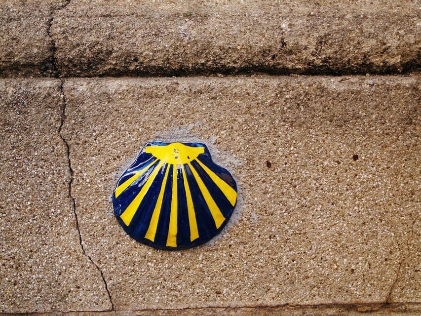 Un primer plano de una concha de vieira de cerámica azul y amarilla, el símbolo del Camino de Santiago, incrustada en una pared de hormigón.