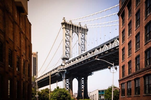Un ponte sospeso in acciaio si erge tra due edifici in mattoni a più piani sotto un cielo sereno.