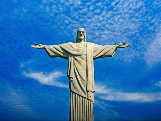 La statua del Cristo Redentore si erge con le braccia aperte contro un cielo blu vibrante punteggiato di nuvole bianche.