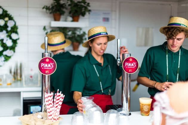 Tre barman in camicia verde e cappelli di paglia servono Pimm's & Lemonade da spillatori presso uno stand per eventi.
