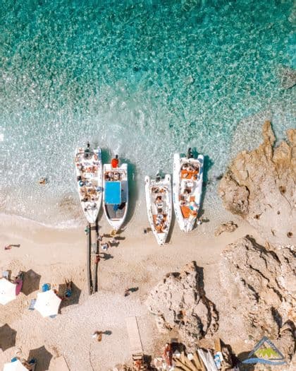 Veduta aerea di un viaggio di gruppo WeRoad con barche ormeggiate su una spiaggia di sabbia, caratterizzata da acque turchesi e cristalline e coste rocciose.
