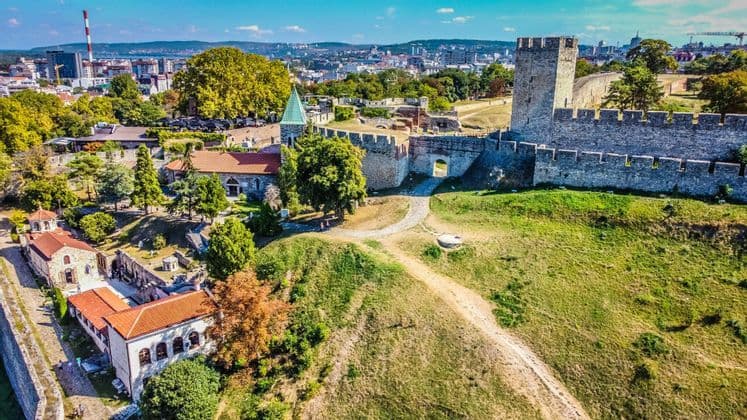 Vista aerea di una storica fortezza in pietra su una collina erbosa, con una chiesa e altri edifici, che domina una città moderna.