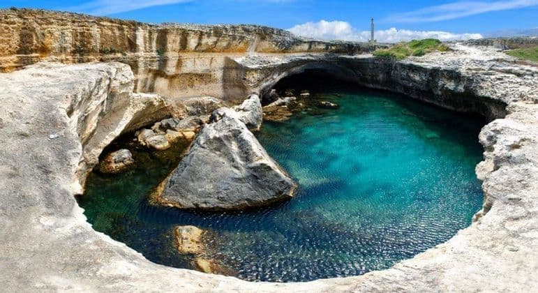 A circular grotto filled with clear turquoise water, surrounded by light-colored rock cliffs under a blue sky.