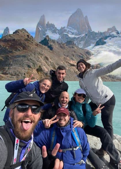 Un grupo de WeRoad se toma una selfie sonriente junto a un lago turquesa con montañas nevadas de fondo.