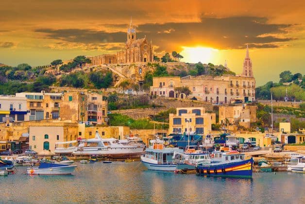 Ein Hafen voller Boote und Yachten vor einer Hangstadt mit einer großen Kirche unter einem goldenen Sonnenuntergang.