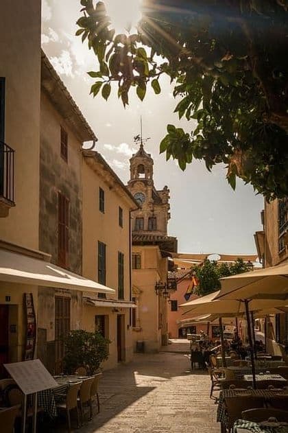 Una calle estrecha y soleada con mesas de café al aire libre, que lleva hacia una torre de reloj visible a través de las ramas de los árboles.