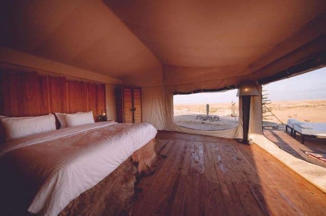 L'interno di una tenda di lusso con un grande letto, pavimenti in legno e un'apertura che rivela un paesaggio desertico.