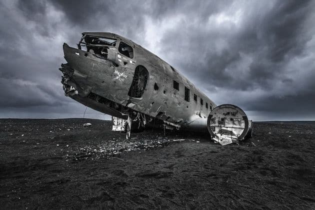 Das Wrack eines Flugzeugrumpfes liegt auf einem Feld aus schwarzem Sand unter einem stark bewölkten Himmel.