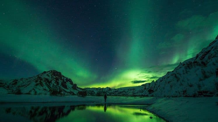Una persona osserva l'Aurora Boreale sopra montagne innevate, il suo bagliore verde che si riflette nell'acqua calma di un lago.