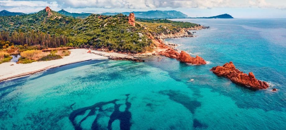 Vista aerea di una costa con acqua turchese, una spiaggia di sabbia bianca, rocce rosse e colline verdi sotto un cielo nuvoloso.