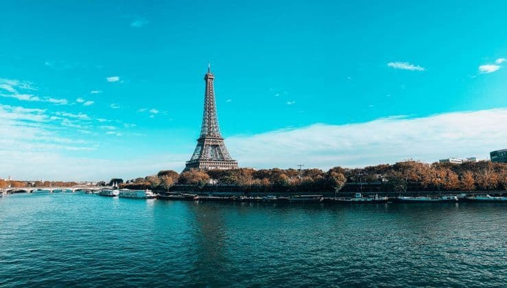 La Torre Eiffel si erge di fronte a un ampio fiume fiancheggiato da alberi autunnali e barche ormeggiate, sotto un cielo azzurro brillante.
