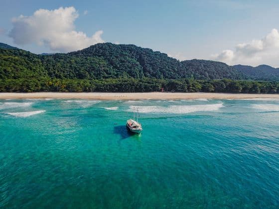 Vue aérienne d'un voilier blanc ancré dans des eaux turquoise, avec une plage de sable et une montagne verdoyante en arrière-plan.