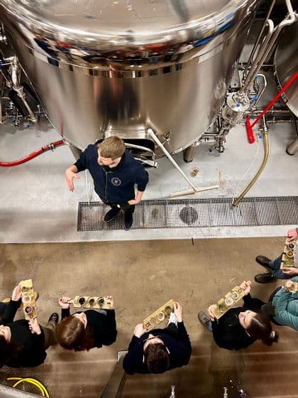 Una guida spiega il processo di birrificazione a un gruppo WeRoad che tiene assaggi di birra accanto a grandi tini d'acciaio in un birrificio.