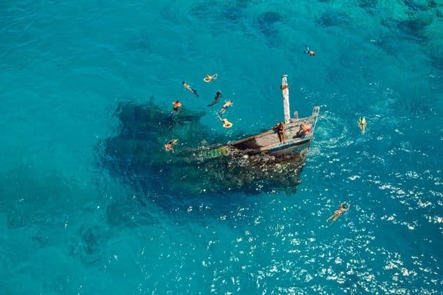 Vue aérienne d'un groupe WeRoad en voyage, faisant de la plongée avec tuba autour d'une épave dans une eau turquoise éclatante.